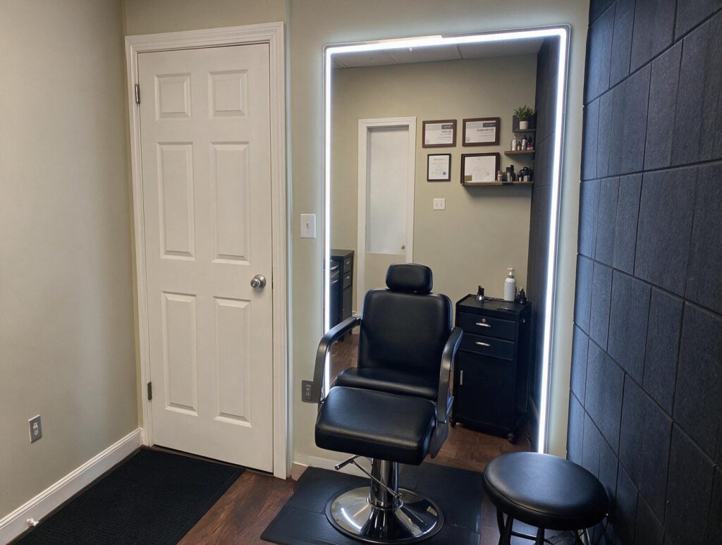 Empty treatment room with a black reclining chair, round stool, lit wall mirror