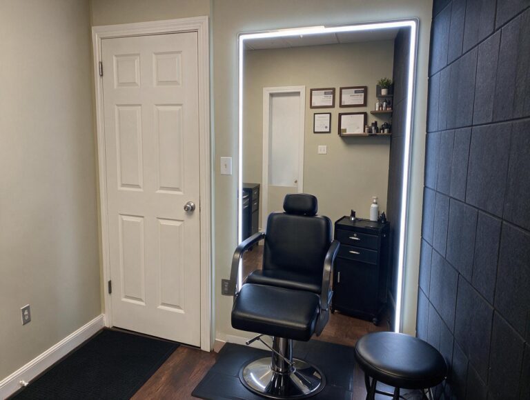 Empty treatment room with a black reclining chair, round stool, lit wall mirror