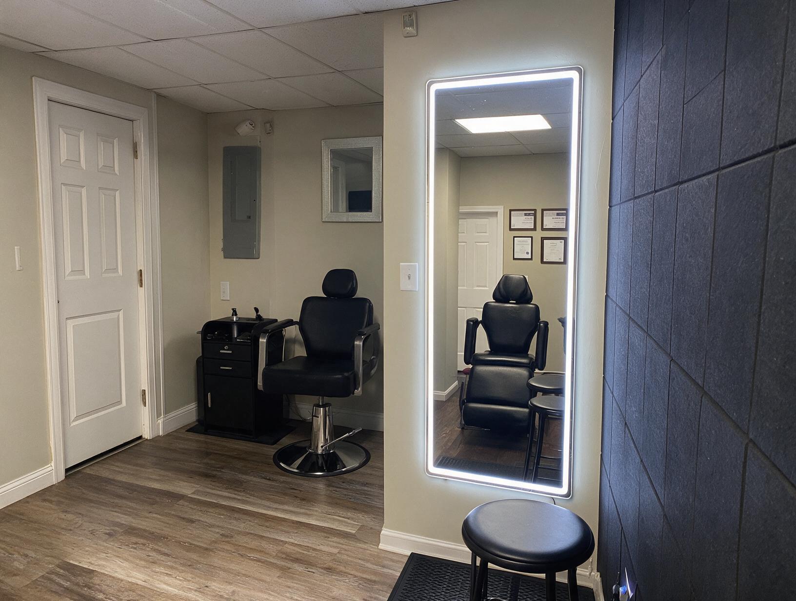 Empty studio room with a black salon chair and cabinet, a lit full-length mirror reflecting a treatment chair
