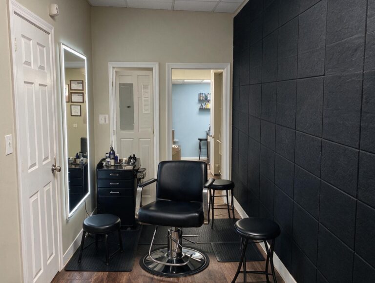 Empty treatment room with a black leather chair, black stools, a lit wall mirror, a black cabinet and a dark paneled wall.