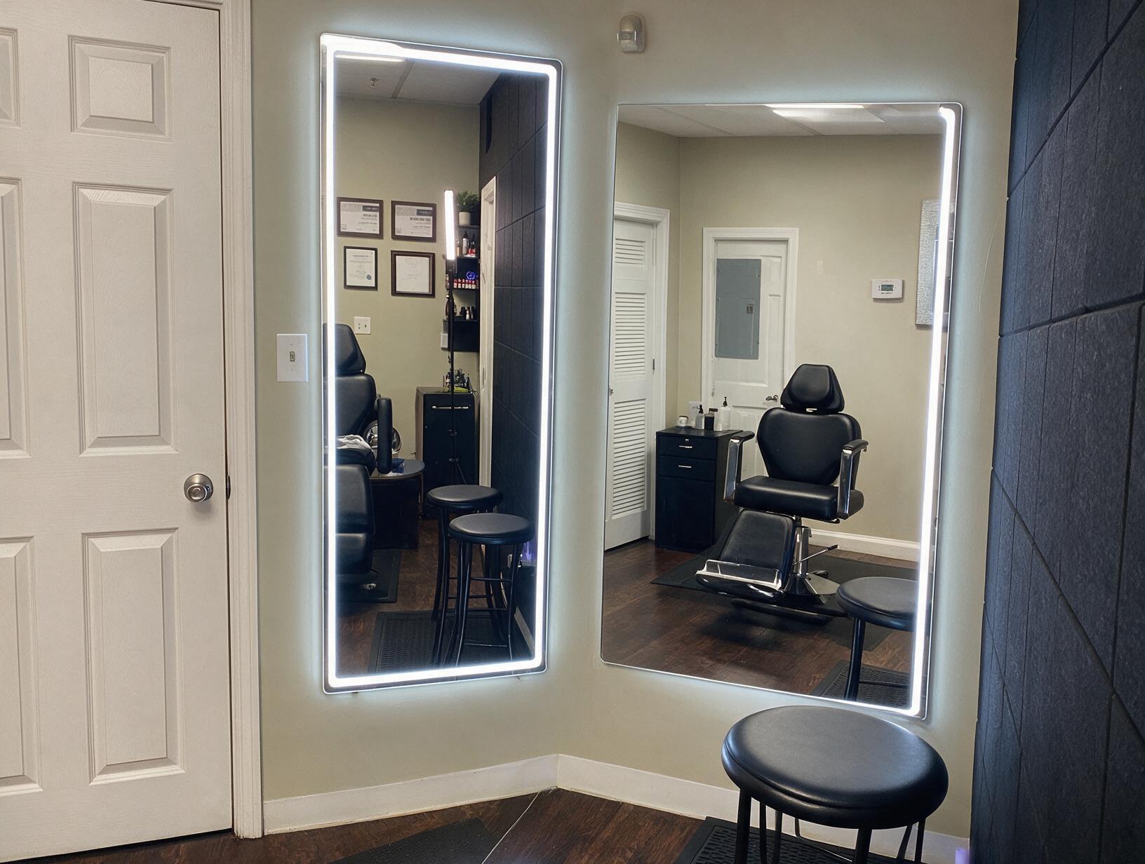 Empty treatment room with two lit wall mirrors, a black reclining chair, black stools, a cabinet and white doors